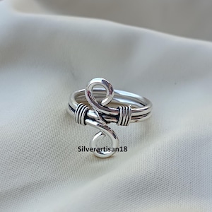 May include: A silver-toned ring with a unique, swirling design. The ring features a double band and an intricate, openwork pattern with a looped element. The design incorporates wrapped wire details. The text "Silverartisan18" is visible.
