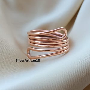 Könnte beinhalten: Ein roségoldfarbener Ring mit einem durchgehenden, mehrschichtigen Spiraldesign aus einem einzigen polierten Metalldraht. Der Ring hat eine offene Wickelstruktur mit glatten, abgerundeten Kanten. Der Text SilverArtisan18 ist zu sehen.