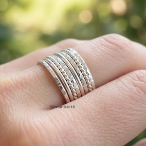 May include: A stack of silver rings with various textures, including hammered and twisted designs. The rings are worn on a finger, showcasing the different styles and finishes. The rings are from Silverartisan18.