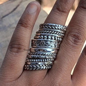 Puede incluir: Una pila de anillos de plata con varios diseños, incluyendo trenzados, con cuentas y florales. Los anillos se llevan en un dedo, mostrando las diferentes texturas y estilos. Los anillos son de metal brillante.