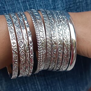 Puede incluir: Una colección de pulseras de plata con intrincados diseños florales y geométricos. Las pulseras están apiladas en una muñeca, mostrando sus diseños detallados y su acabado pulido. Las pulseras son de plata.