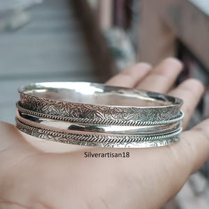Könnte beinhalten: Ein silbernes Armreif-Armband mit aufwendigen floralen und Seildetails. Das Armband wird in einer Hand gehalten und zeigt seine polierte Oberfläche und gravierten Muster. Ein klassisches Schmuckstück.