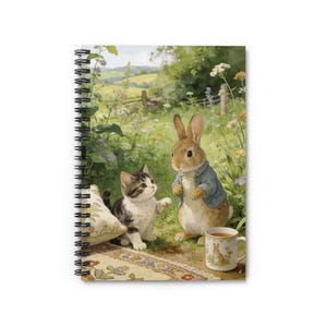 Può includere: Quaderno a spirale con un'illustrazione ad acquerello di un gattino e un coniglio in un campo. Il coniglio indossa una giacca blu e una tazza da tè con un disegno di coniglio è vicina. La scena è ambientata in un prato verde lussureggiante.