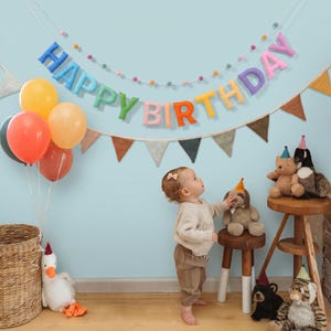 Può includere: Uno striscione in feltro Happy Birthday con lettere multicolori e una ghirlanda di pon pon, un grappolo di palloncini arancioni e gialli, un cesto di vimini, un'oca di peluche bianca, un bambino con maglione beige e vari peluche su sgabelli.