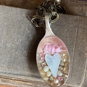 May include: A silver spoon necklace with a pink pig figurine, a white heart with a blue floral design, and white and pink beads.