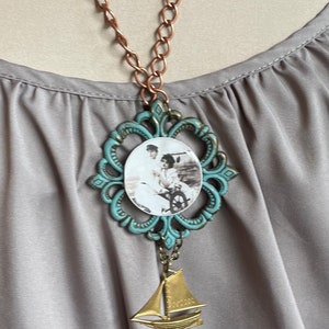 May include: A necklace with a turquoise blue ornate frame pendant featuring a vintage photograph of a couple. The pendant is attached to a copper chain and a gold sailboat charm.