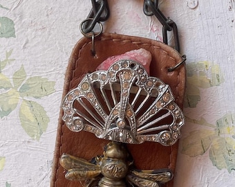 May include: A handmade pendant necklace featuring a brown leather backing with a silver fan-shaped embellishment and a brass bee. A small white rectangle with the word "rock" is attached. The necklace has a chain.