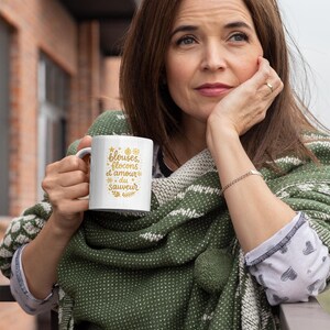 May include: A white ceramic mug with the text "blouses, flocons et amour du sauveur" in gold lettering. The mug is held by a person wearing a green shawl with a white pattern. The person is outside, leaning on a railing.