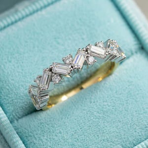 May include: A white gold ring featuring a zigzag design of baguette and round cut diamonds. The ring is displayed against a light blue backdrop, highlighting the sparkle of the gemstones. The band has a gold interior.