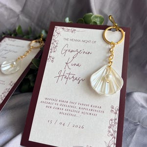 以下が含まれることがあります： 真珠のアクセントが付いた貝殻型のチャームが付いた結婚式の記念品。ゴールドのキーリングに付いています。チャームは、トルコ語のテキストと日付15/04/2026が記載された、バーガンディとクリーム色のカードと組み合わされています。