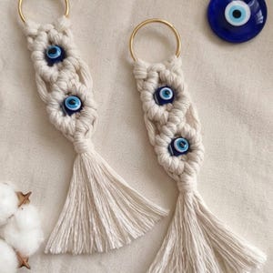 May include: Two macrame keychains made of off-white cord, each featuring two blue and white evil eye beads and a gold-toned metal ring. A separate circular blue and white evil eye glass charm is positioned near the top right of the keychains.
