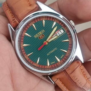 K&ouml;nnte beinhalten: Vintage Seiko 5 Automatik-Uhr mit gr&uuml;nem Zifferblatt und braunem Lederarmband. Das Zifferblatt der Uhr hat goldfarbene Stundenmarkierungen und Zeiger mit einem roten Sekundenzeiger. Die Uhr hat ein Edelstahlgeh&auml;use und eine Datumsanzeige bei 3 Uhr.