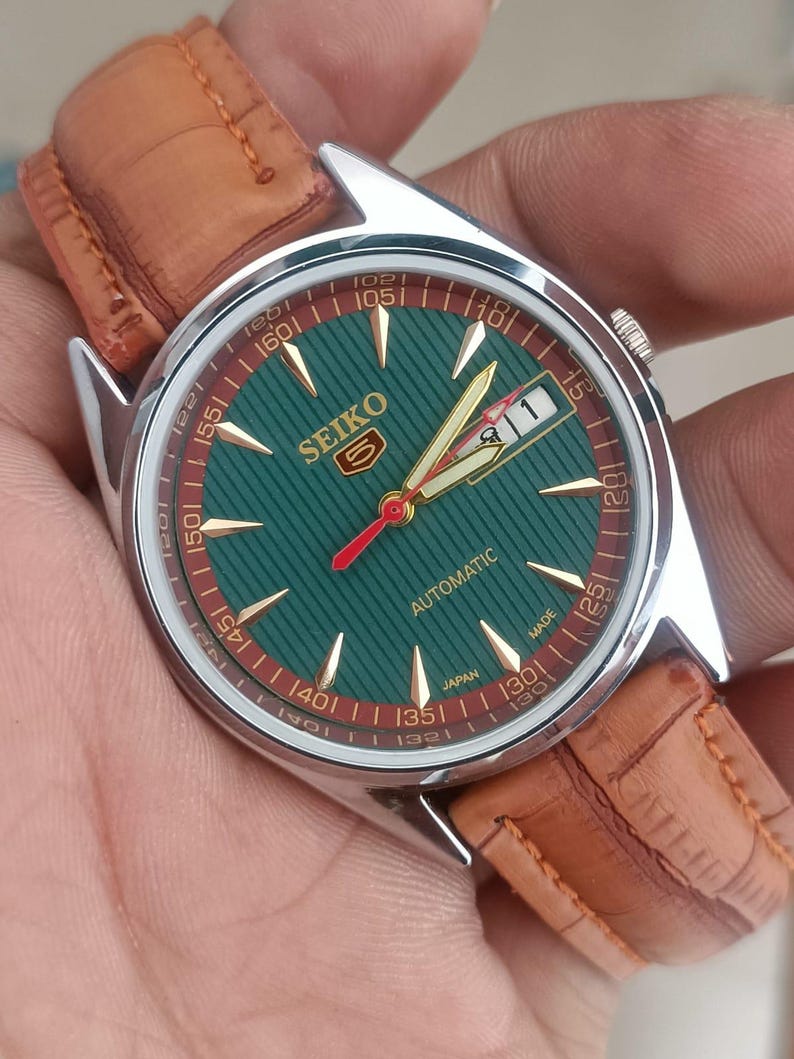K&ouml;nnte beinhalten: Vintage Seiko 5 Automatik-Uhr mit gr&uuml;nem Zifferblatt und braunem Lederarmband. Das Zifferblatt der Uhr hat goldfarbene Stundenmarkierungen und Zeiger mit einem roten Sekundenzeiger. Die Uhr hat ein Edelstahlgeh&auml;use und eine Datumsanzeige bei 3 Uhr.