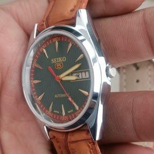 K&ouml;nnte beinhalten: Eine silberfarbene Seiko Automatik-Armbanduhr mit dunkelgr&uuml;nem Zifferblatt, goldfarbenen Zeigern und Markierungen sowie Datumsanzeige. Sie hat ein braunes Lederarmband. Auf dem Zifferblatt steht "SEIKO" und die Zahl "5".