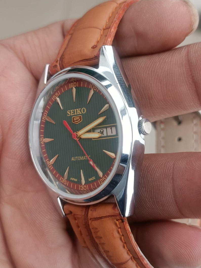 K&ouml;nnte beinhalten: Eine silberfarbene Seiko Automatik-Armbanduhr mit dunkelgr&uuml;nem Zifferblatt, goldfarbenen Zeigern und Markierungen sowie Datumsanzeige. Sie hat ein braunes Lederarmband. Auf dem Zifferblatt steht "SEIKO" und die Zahl "5".