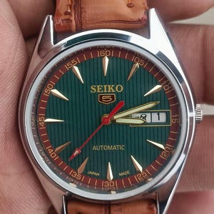 K&ouml;nnte beinhalten: Eine Vintage Seiko Automatik-Armbanduhr mit gr&uuml;nem Zifferblatt und goldenen Akzenten. Die Uhr verf&uuml;gt &uuml;ber ein braunes Lederarmband und eine Datumsanzeige. Das Zifferblatt hat ein vertikal gestreiftes Muster und goldene Zeiger.
