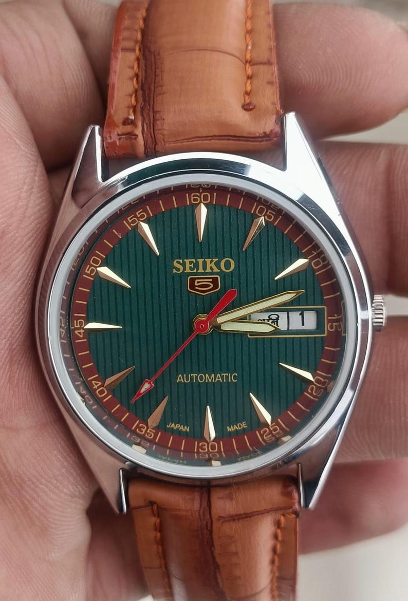 K&ouml;nnte beinhalten: Eine Vintage Seiko Automatik-Armbanduhr mit gr&uuml;nem Zifferblatt und goldenen Akzenten. Die Uhr verf&uuml;gt &uuml;ber ein braunes Lederarmband und eine Datumsanzeige. Das Zifferblatt hat ein vertikal gestreiftes Muster und goldene Zeiger.