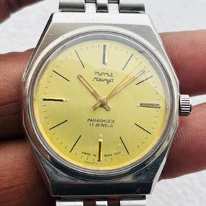 Op de afbeelding: Een vintage polshorloge met een zilverkleurige metalen band en kast. De wijzerplaat is lichtgeel met zwarte markeringen en wijzers. De wijzerplaat heeft de tekst "Rumu Maurya" en "Parashock 17 Jewels".