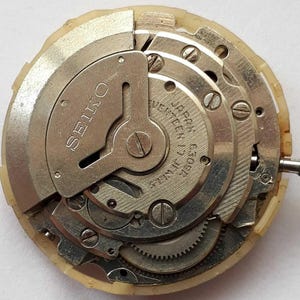 K&ouml;nnte beinhalten: Nahaufnahme eines Seiko-Uhrwerks. Die Metallkomponenten sind silber- und goldfarben, mit sichtbaren Zahnr&auml;dern, Schrauben und dem Text "SEIKO" und "JAPAN 17 JEWELS C309A". Das Uhrwerk ist in einem cremefarbenen Kunststoffgeh&auml;use untergebracht.