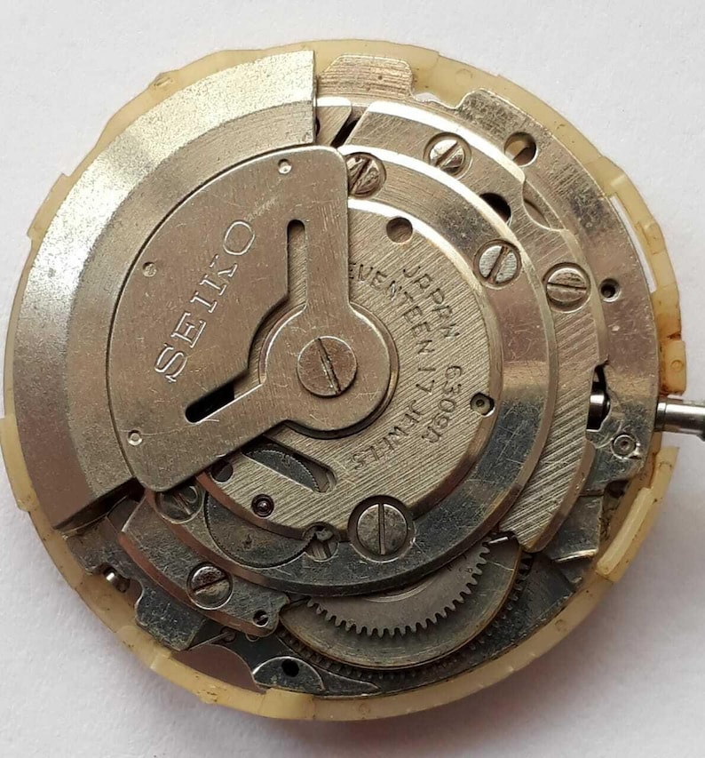 K&ouml;nnte beinhalten: Nahaufnahme eines Seiko-Uhrwerks. Die Metallkomponenten sind silber- und goldfarben, mit sichtbaren Zahnr&auml;dern, Schrauben und dem Text "SEIKO" und "JAPAN 17 JEWELS C309A". Das Uhrwerk ist in einem cremefarbenen Kunststoffgeh&auml;use untergebracht.