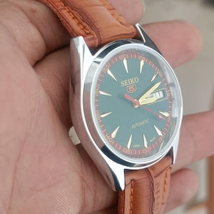 K&ouml;nnte beinhalten: Seiko Automatik-Uhr mit silbernem Geh&auml;use, gr&uuml;nem Zifferblatt und braunem Lederarmband. Die Uhr hat goldene Zeiger und zeigt Tag und Datum an. Das Armband ist braun mit sichtbaren N&auml;hten.
