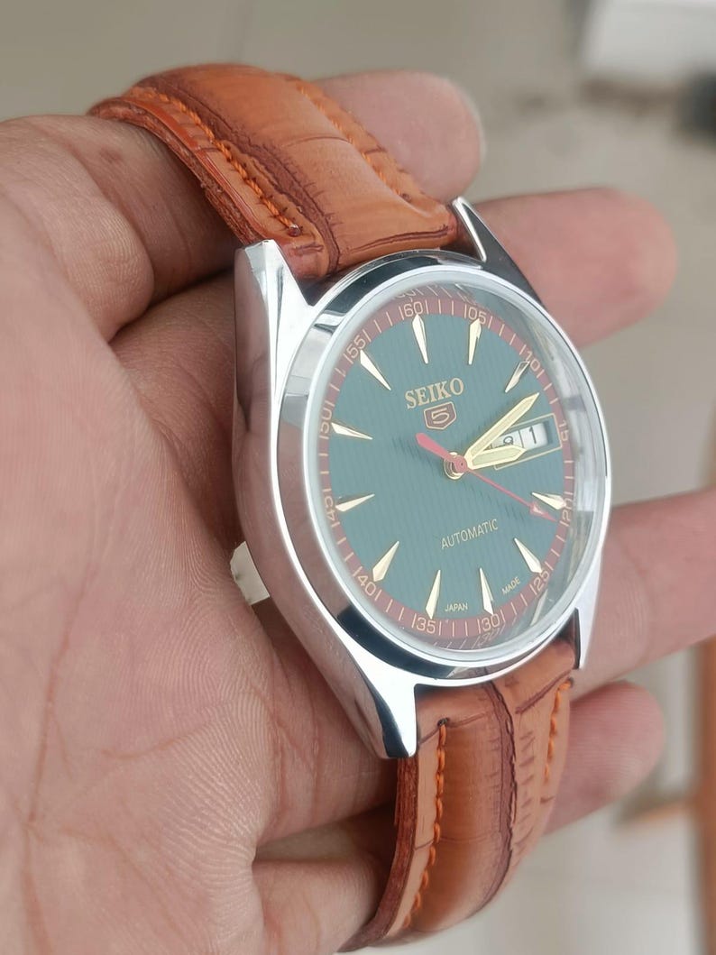 K&ouml;nnte beinhalten: Seiko Automatik-Uhr mit silbernem Geh&auml;use, gr&uuml;nem Zifferblatt und braunem Lederarmband. Die Uhr hat goldene Zeiger und zeigt Tag und Datum an. Das Armband ist braun mit sichtbaren N&auml;hten.