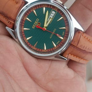K&ouml;nnte beinhalten: Seiko Automatik-Armbanduhr mit gr&uuml;nem Zifferblatt und goldenen Zeigern. Die Uhr hat ein silbernes Geh&auml;use und ein braunes Lederarmband. Das Zifferblatt hat goldene Markierungen und ein Datumsfenster bei 3 Uhr.