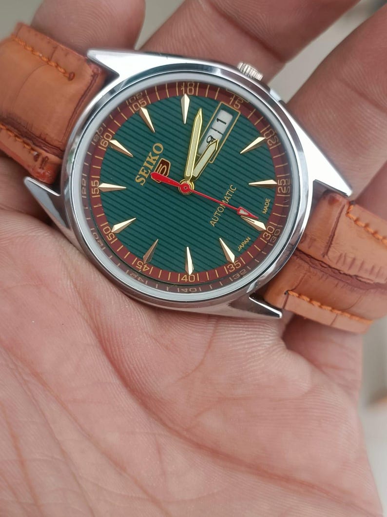 K&ouml;nnte beinhalten: Seiko Automatik-Armbanduhr mit gr&uuml;nem Zifferblatt und goldenen Zeigern. Die Uhr hat ein silbernes Geh&auml;use und ein braunes Lederarmband. Das Zifferblatt hat goldene Markierungen und ein Datumsfenster bei 3 Uhr.