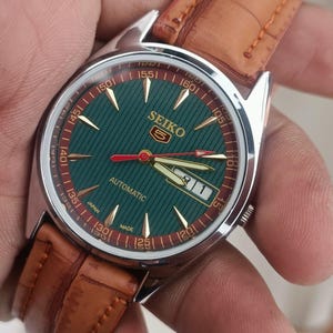 K&ouml;nnte beinhalten: Eine Seiko Automatik-Armbanduhr mit gr&uuml;nem Zifferblatt und goldfarbenen Zeigern und Markierungen. Die Uhr hat ein braunes Lederarmband und ein Edelstahlgeh&auml;use. Das Zifferblatt tr&auml;gt die Aufschrift "SEIKO" und "AUTOMATIC".