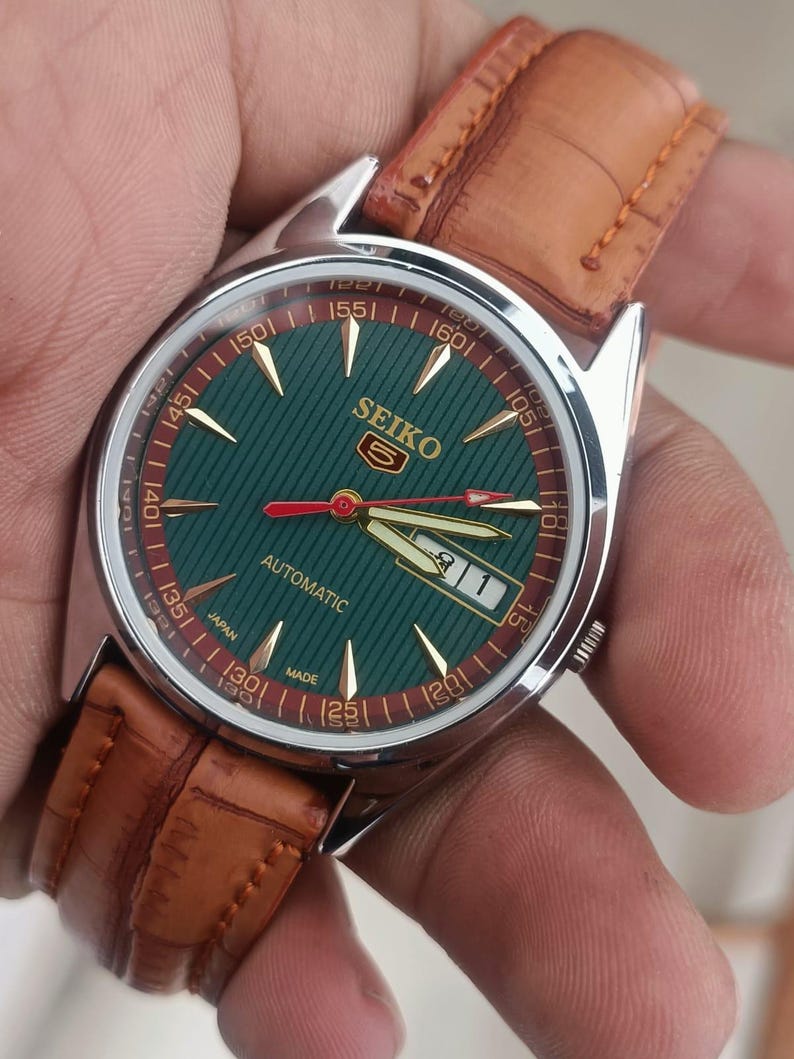 K&ouml;nnte beinhalten: Eine Seiko Automatik-Armbanduhr mit gr&uuml;nem Zifferblatt und goldfarbenen Zeigern und Markierungen. Die Uhr hat ein braunes Lederarmband und ein Edelstahlgeh&auml;use. Das Zifferblatt tr&auml;gt die Aufschrift "SEIKO" und "AUTOMATIC".