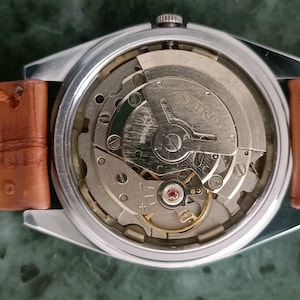K&ouml;nnte beinhalten: Nahaufnahme einer Vintage Seiko Uhr, die das komplizierte mechanische Uhrwerk zeigt. Die Uhr hat ein silberfarbenes Geh&auml;use und ein braunes Lederarmband. Die inneren Zahnr&auml;der und Komponenten sind sichtbar, mit dem Seiko-Logo.