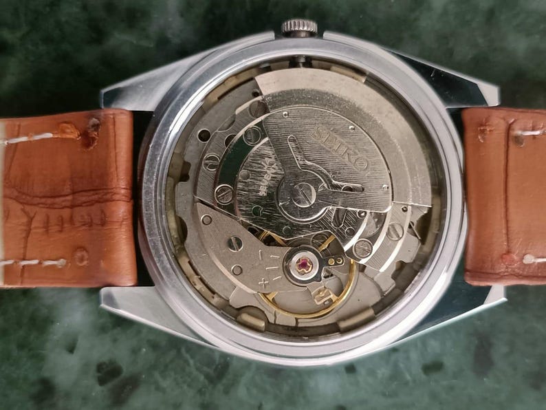 K&ouml;nnte beinhalten: Nahaufnahme einer Vintage Seiko Uhr, die das komplizierte mechanische Uhrwerk zeigt. Die Uhr hat ein silberfarbenes Geh&auml;use und ein braunes Lederarmband. Die inneren Zahnr&auml;der und Komponenten sind sichtbar, mit dem Seiko-Logo.