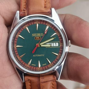 K&ouml;nnte beinhalten: Eine Vintage Seiko Automatik-Armbanduhr mit gr&uuml;nem Zifferblatt und braunem Lederarmband. Das Zifferblatt hat goldfarbene Zeiger und Markierungen, einen roten Sekundenzeiger und eine Datumsanzeige. Das Uhrengeh&auml;use ist silberfarben.