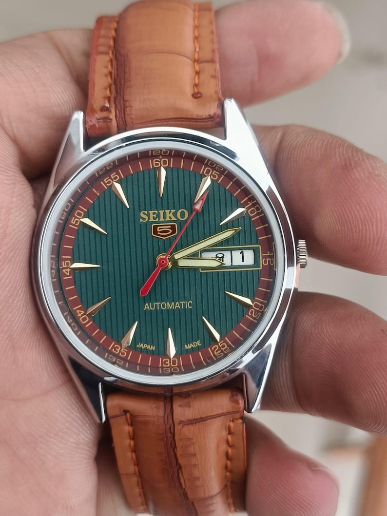 K&ouml;nnte beinhalten: Eine Vintage Seiko Automatik-Armbanduhr mit gr&uuml;nem Zifferblatt und braunem Lederarmband. Das Zifferblatt hat goldfarbene Zeiger und Markierungen, einen roten Sekundenzeiger und eine Datumsanzeige. Das Uhrengeh&auml;use ist silberfarben.