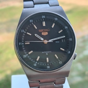 Pode incluir: Um relógio de pulso automático Seiko vintage com mostrador preto e ponteiros e marcadores prateados. O relógio apresenta uma pulseira de aço inoxidável e um visor de data. O mostrador exibe o logotipo Seiko e a palavra "AUTOMATIC".