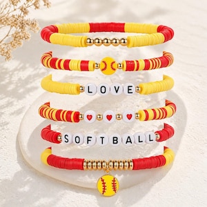 Puede incluir: Una colección de pulseras con temática de softbol en rojo, amarillo y dorado. Algunas pulseras presentan las palabras "LOVE" y "SOFTBALL" deletreadas con cuentas de letras blancas. Otras pulseras tienen cuentas en forma de corazón o un dije de softbol.