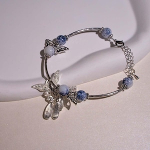 May include: A silver bracelet featuring butterfly charms and blue and white speckled beads. The bracelet has a curved design with a chain extension for adjustable sizing. The butterfly charms are intricately detailed.