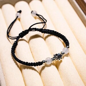 May include: Black braided bracelet with white beads and a silver charm. The adjustable bracelet has a woven design with a sliding knot closure. The charm has a floral design. The bracelet is displayed on a cream-coloured surface.