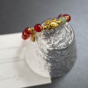 May include: A gold-colored bracelet with red and green beads. The bracelet features a gold charm with a detailed design. The beads are round and evenly spaced. The bracelet is displayed on a textured silver surface.