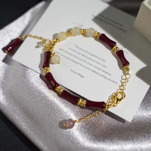 May include: A gold-colored chain bracelet with alternating burgundy bamboo-shaped beads and cream-colored round beads. The bracelet includes a small charm and a clasp. A card with text is in the background.
