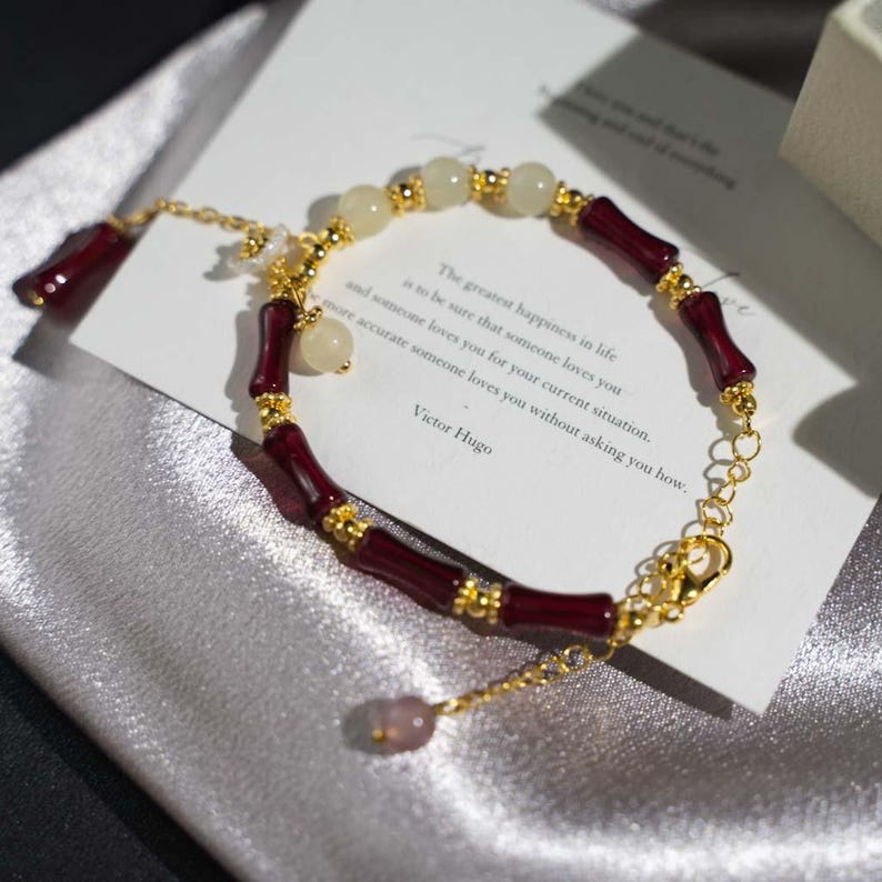 May include: A gold-colored chain bracelet with alternating burgundy bamboo-shaped beads and cream-colored round beads. The bracelet includes a small charm and a clasp. A card with text is in the background.