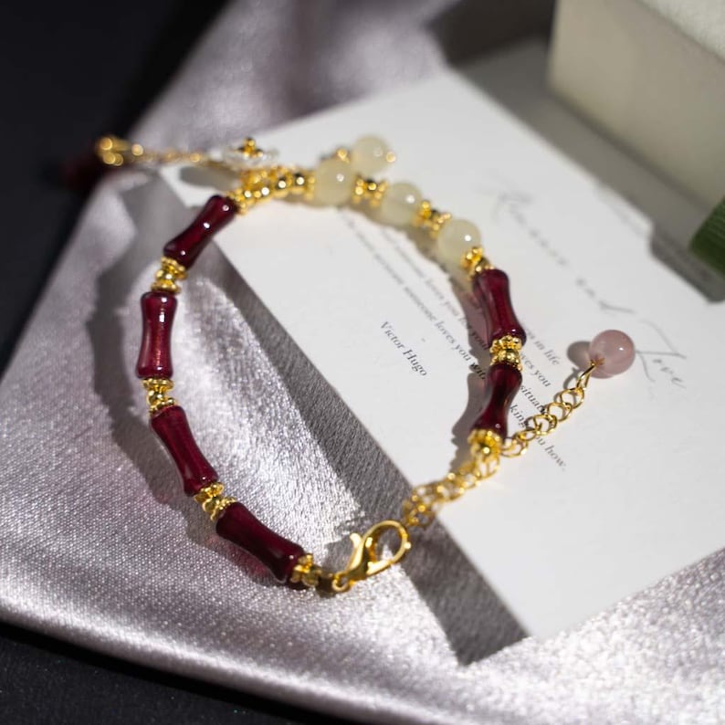 May include: A gold-toned bracelet featuring alternating segments of dark red bamboo-shaped beads and light-colored, round beads. The bracelet includes a delicate chain with a pink gemstone charm and a clasp closure.