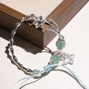 Puede incluir: Pulsera plateada con un diseño de bambú y elementos decorativos. Incluye un detalle floral, dos cuentas de piedra verde y un dije de mariposa. La pulsera se asegura con un cordón azul claro.