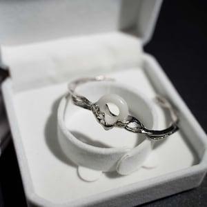 May include: A silver-toned bracelet with a white circular stone is displayed inside a white jewelry box. The bracelet has a twisted design and a smooth, polished finish. The box is open, revealing a soft, white interior.