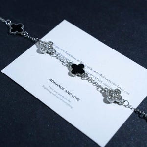 Könnte beinhalten: Ein silbernes Armband mit Kleeblatt-Anhängern. Einige sind schwarz, andere mit kleinen, klaren Steinen besetzt. Das Armband liegt auf einer weißen Karte mit der Aufschrift "ROMANCE AND LOVE".