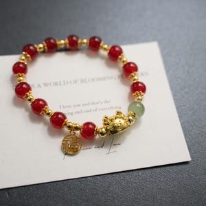 May include: A beaded bracelet featuring red and gold beads, with a gold charm and a green bead. The bracelet is on a white card with text that reads "A WORLD OF BLOOMING FLOWERS".