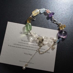 Puede incluir: Una pulsera de cuentas con una variedad de piedras preciosas coloridas, incluyendo piedras amarillas, verdes, claras y moradas. La pulsera está conectada con una cadena y un cierre plateados. Una tarjeta con la palabra "Romance" y una cita de Victor Hugo está en el fondo.