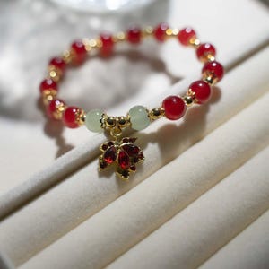 May include: A beaded bracelet featuring red and gold beads, with two light green beads and a gold charm shaped like a leaf with red accents. The bracelet is displayed on a white surface.