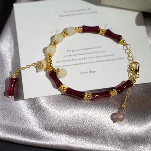 May include: A gold-toned bracelet with burgundy bamboo-shaped beads and cream-colored spherical beads. The bracelet includes a small flower charm and a dangling burgundy bead. A card with text by Victor Hugo is in the background.