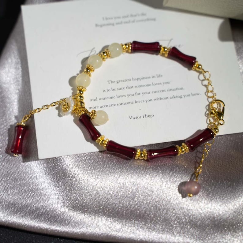 May include: A gold-toned bracelet with burgundy bamboo-shaped beads and cream-colored spherical beads. The bracelet includes a small flower charm and a dangling burgundy bead. A card with text by Victor Hugo is in the background.